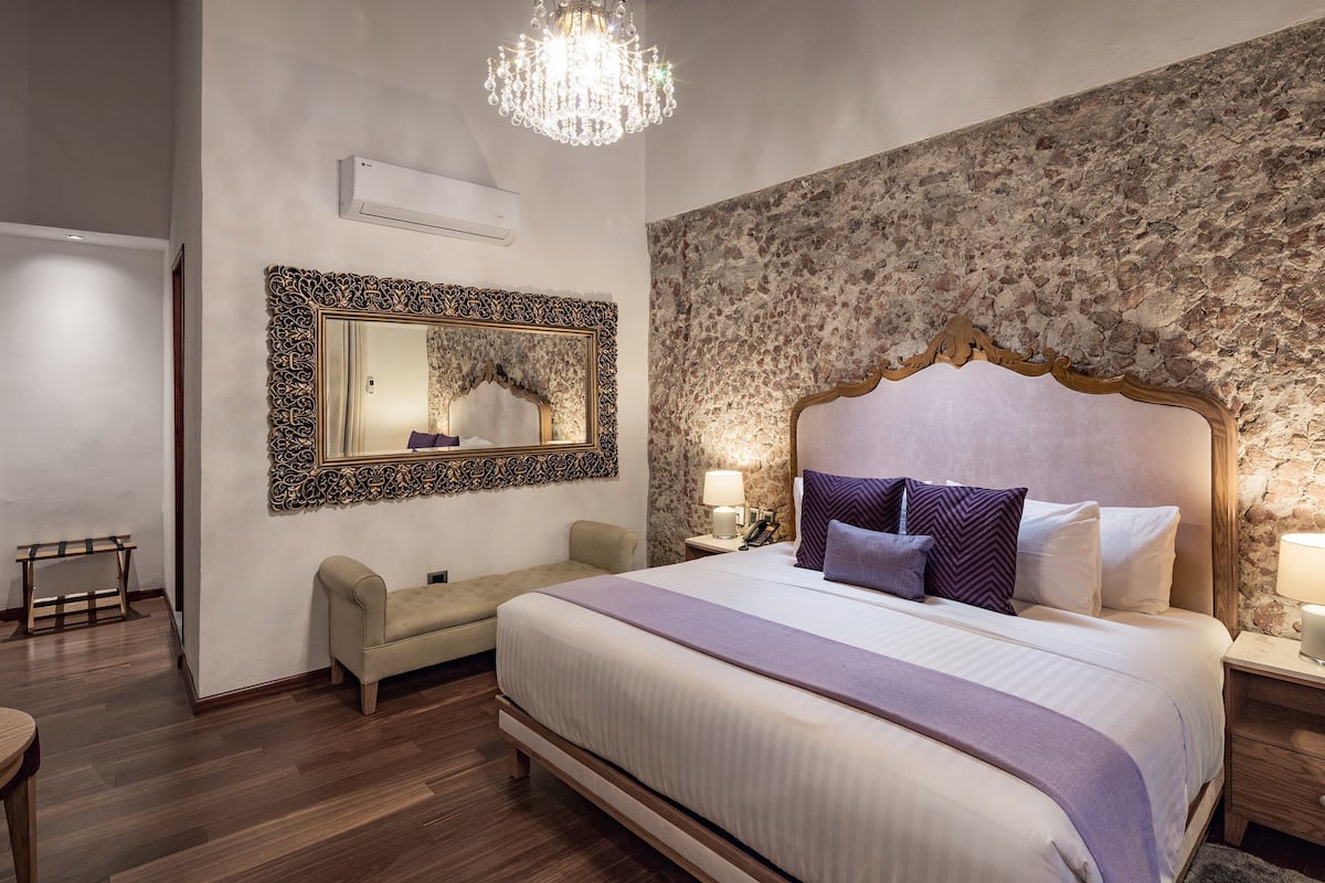 A hotel room with a large bed, a nightstand, a bedside table with a lamp, a chair, a bench, a chandelier, and a stone wall.