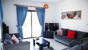 A living room with a sofa, coffee table, and TV. There are blue curtains and a painting on the wall. - Stunning 4 Bedroom Villa -Private Pool - Sea View (Peyia)