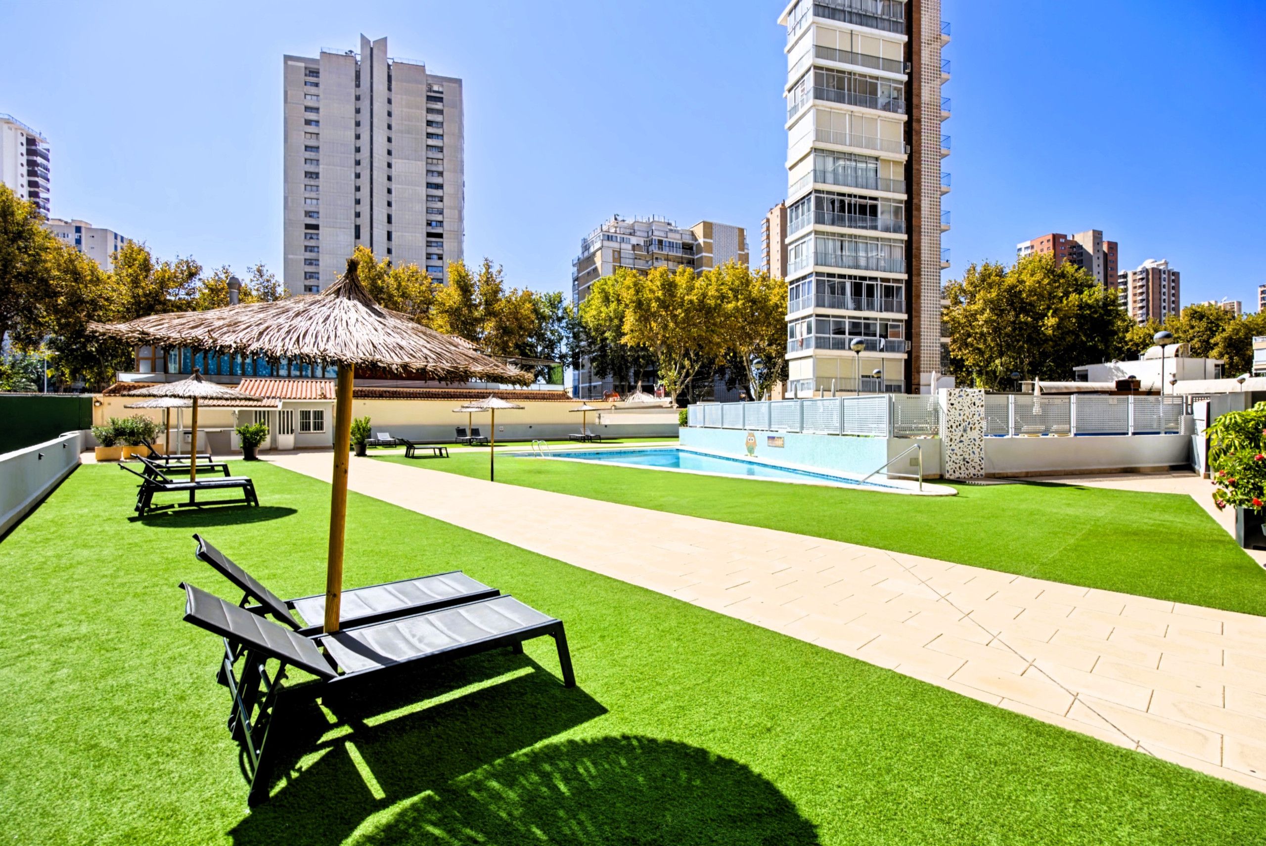 A rooftop pool area with artificial grass, lounge chairs, and a thatched umbrella.
