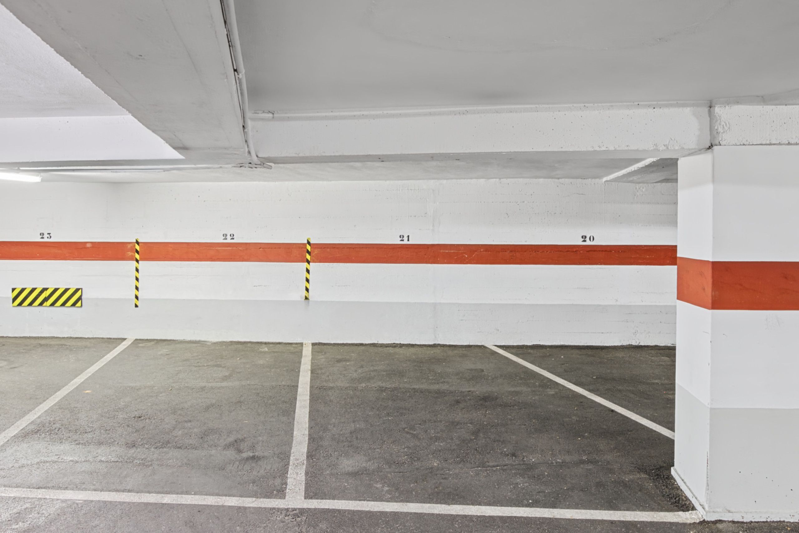 A parking garage with white walls and red stripes, numbered parking spots, and yellow and black striped bollards.