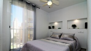 A bedroom with a bed, a ceiling fan, a window with curtains, and wall-mounted shelves with plants. - Ocean II 9-C (Benidorm)