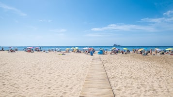 En sandstrand med en träpromenad som leder ner till vattnet, parasoller och människor som kopplar av.