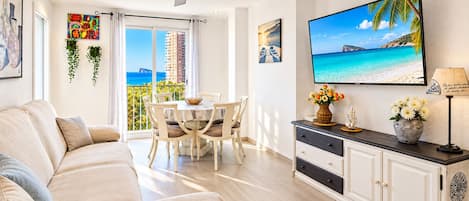A modern living room with a sofa, dining table, and chairs, a ceiling fan, and a TV displaying a beach scene.