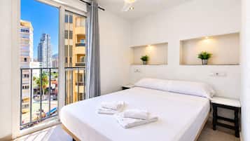 Ett hotellrum med en säng, ett nattduksbord, en vägghylla med en växt, en takfläkt och utsikt över en stadssiluett genom ett stort fönster.