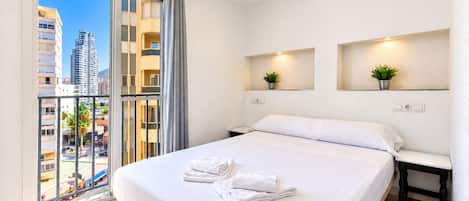 A hotel room with a bed, a nightstand, a wall-mounted shelf with a plant, a ceiling fan, and a view of a cityscape through a large window.