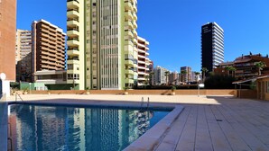 A swimming pool with a tiled deck, surrounded by high-rise buildings and a clear blue sky. - Ocean II 9-C (Benidorm)