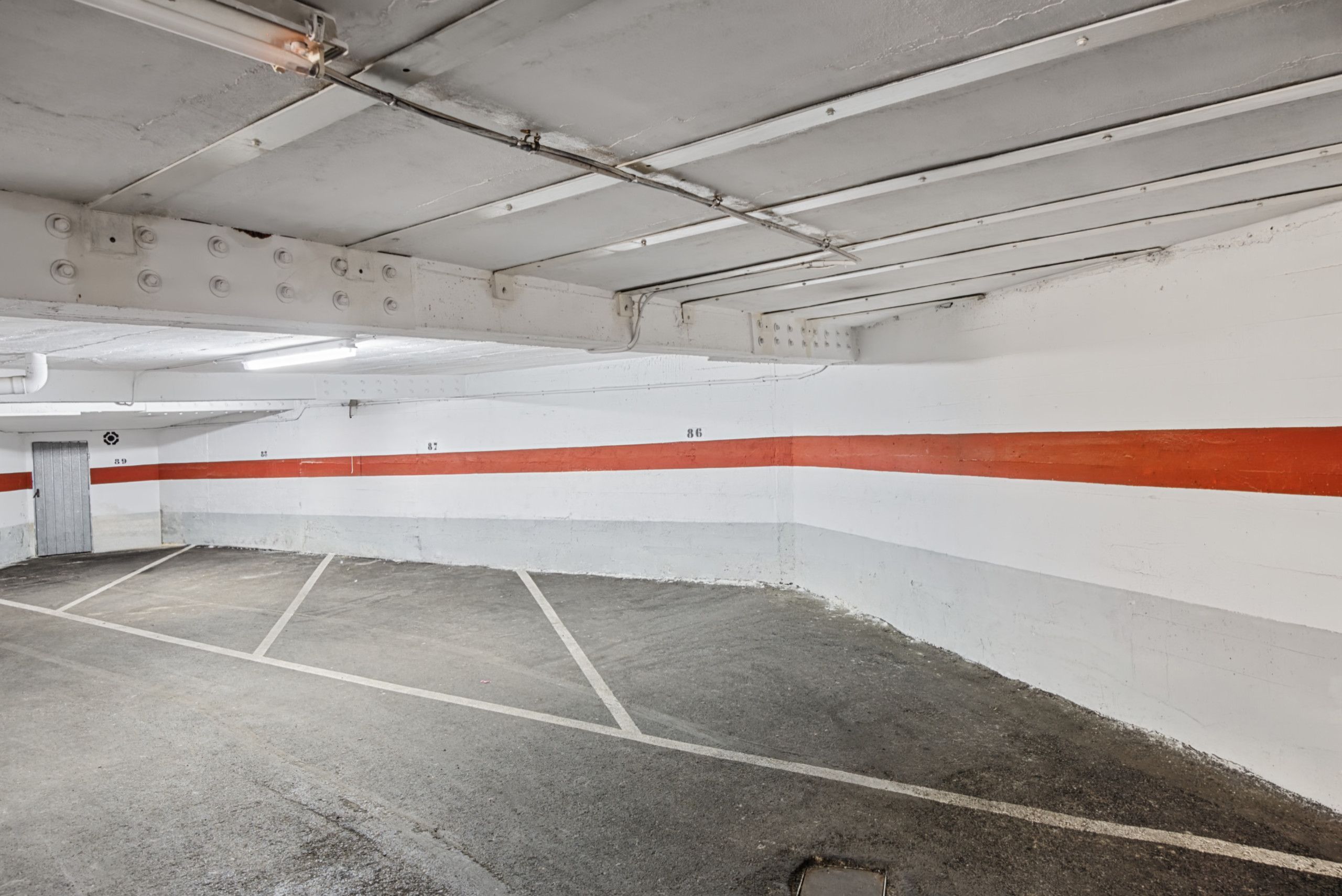 An indoor parking garage with concrete floors and walls, marked parking spaces, and a red and white stripe along the wall.