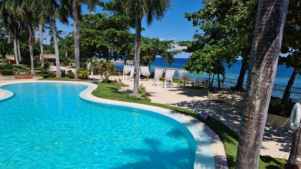 A clear blue pool with a curved edge, surrounded by a sandy beach and palm trees. - Lux. 2BR condo + 2 pools, woow oceanviews, Gym, Beach,near Mactan Newtown, Tours (Lapu-Lapu City)