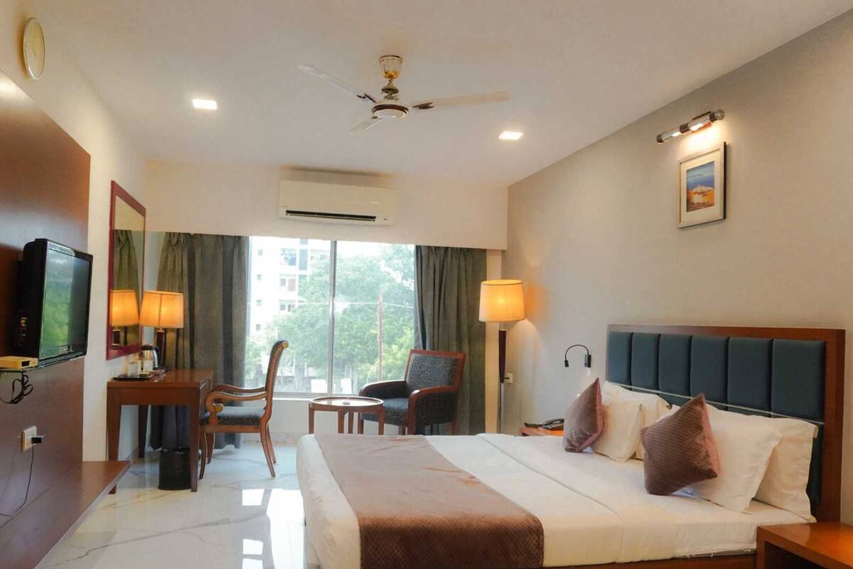 A hotel room with a bed, a desk with a television, two chairs, a lamp, and a ceiling fan.