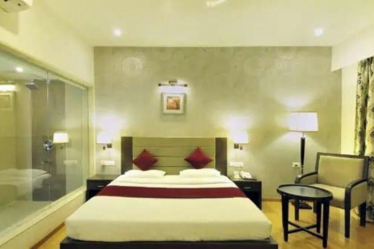 A hotel room with a bed, a bedside table, a chair, a small table, and a glass-enclosed shower area.