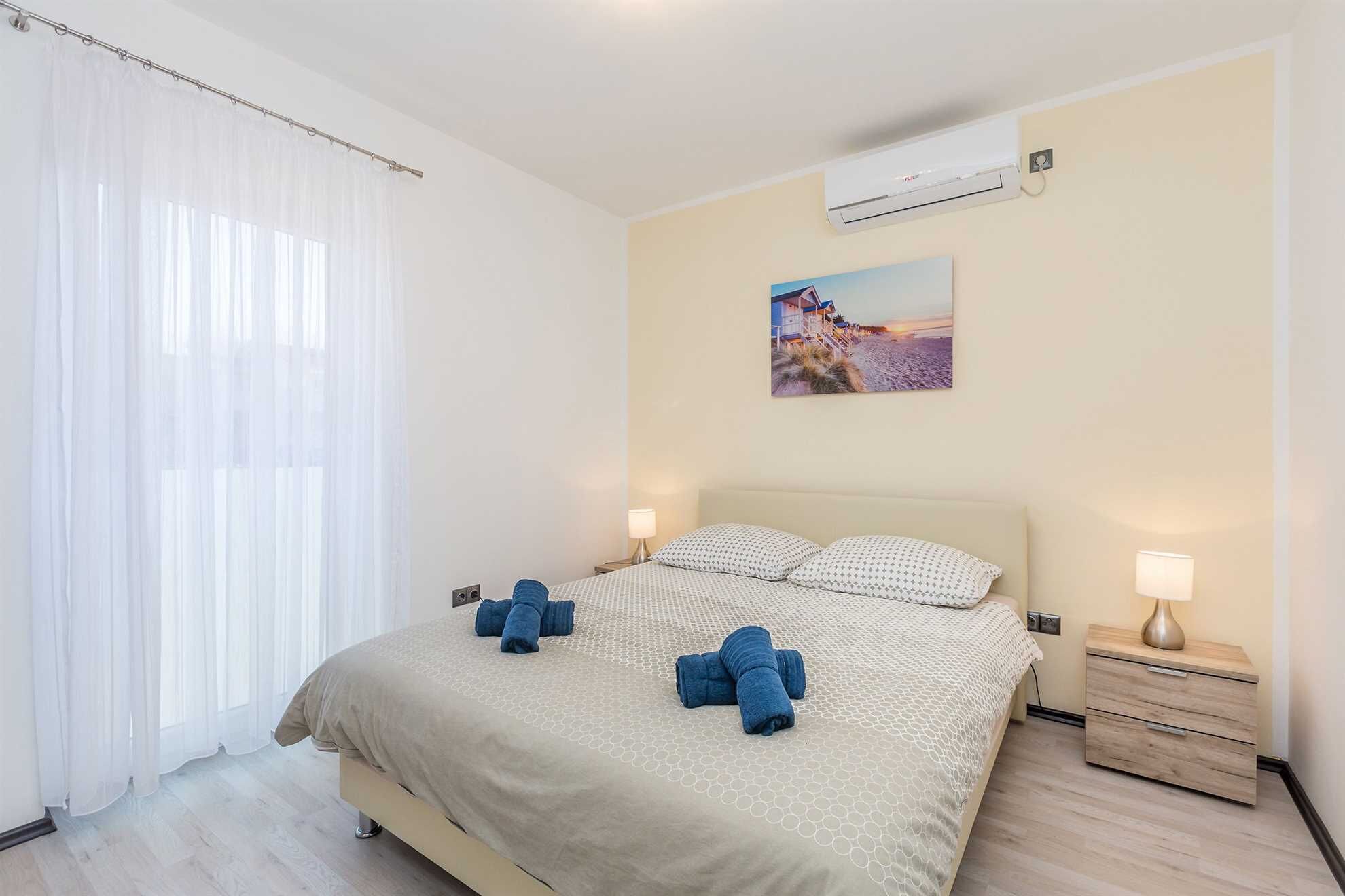 A hotel room with a bed, two bedside tables, an air conditioning unit, and a painting on the wall.