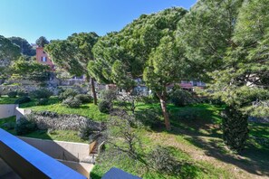 A landscaped garden with various trees and shrubs, a stone wall, and a building in the background. - LOVELY STUDIO 1 ROOM- between Monaco and Menton (Roquebrune-Cap-Martin)