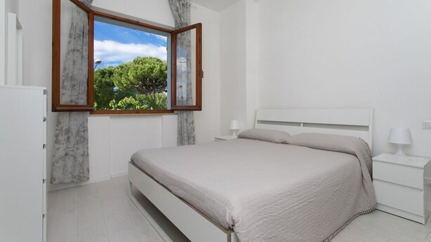 Ein Schlafzimmer mit einem Bett, einem Nachttisch, einem Fenster mit Vorhängen und Blick auf Grün.
