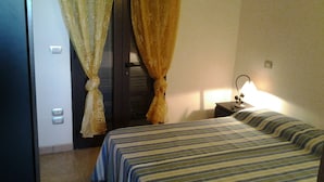 Quarto de hotel com duas camas, mesa de cabeceira com abajur e cortinas.