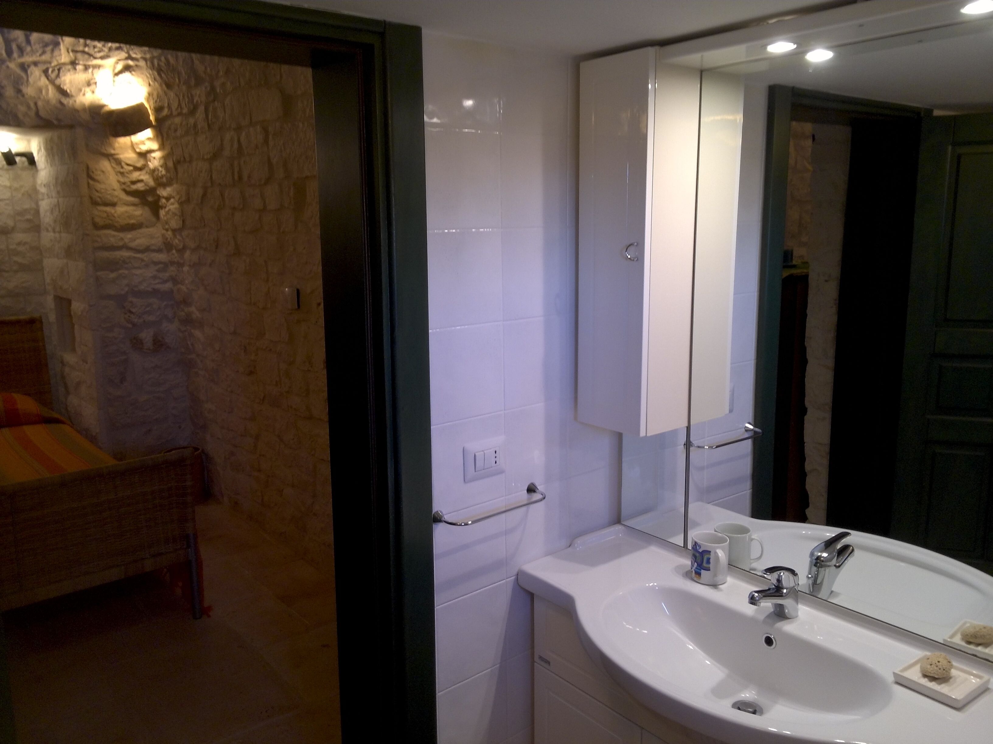 A bathroom with a white sink, mirror, and towel rack. There is a bed with a wooden headboard in the adjacent room.