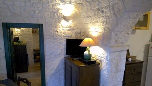 Ein Zimmer mit Steinwänden, einem Fernseher auf einer Holzkommode, einer Lampe und einem kleinen Tisch mit Stuhl.