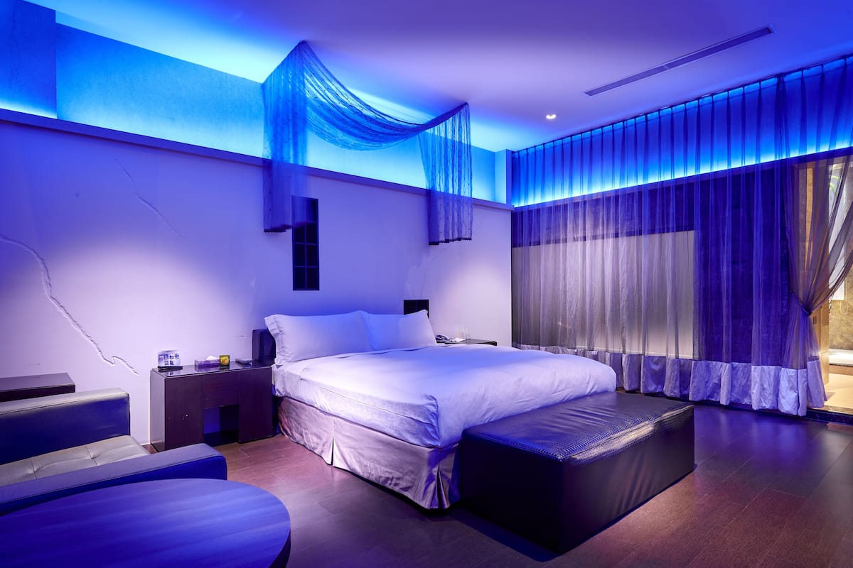 Quarto moderno com cama grande, mesa de cabeceira, sofá e janela com cortinas leves.