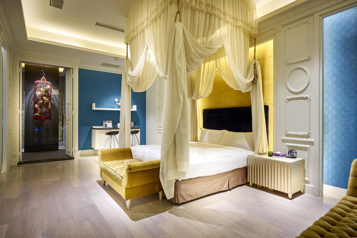 Um quarto luxuoso com uma cama de dossel, um espaço de estar com um sofá amarelo e uma mesa de cabeceira com abajur.