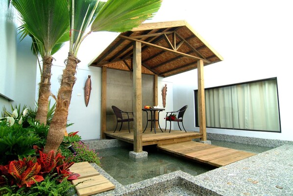Un pabellón de madera con techo de paja, dos sillas y una mesa en un jardín con plantas y suelo de hormigón.