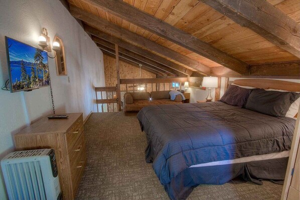 A cozy attic bedroom with a wooden ceiling, a large bed, a nightstand, a dresser, and a painting of a forest scene. - Tahoe Olympic Condo | Carson Valley Views (Stateline)