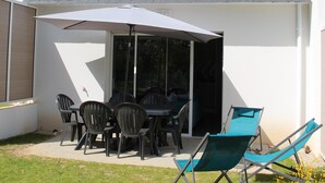 Une maison dotée d’un patio comprenant une table et des chaises, un grand parasol et deux fauteuils de jardin.