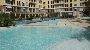 Zona de piscina en un resort, con agua cristalina, rodeada por un edificio con balcones y un paseo techado.