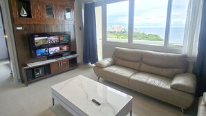 Uma sala de estar moderna com um sofá de couro, uma mesa de centro branca e uma TV em um rack de madeira. Grandes janelas oferecem vista para a paisagem urbana.