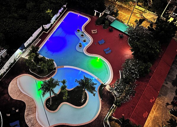 An aerial view of a swimming pool with a wavy edge design, surrounded by a red deck area and palm trees.
