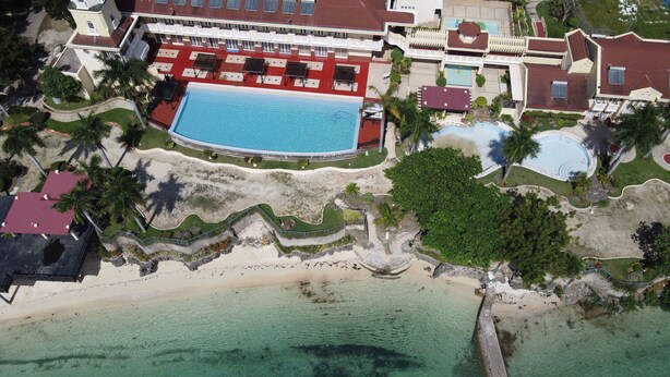 Vista aérea de um resort com uma grande piscina, frente para a praia e vários edifícios com telhados vermelhos.