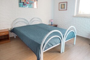 A bed with a white metal frame and a teal bedspread in a room with wooden flooring, a small wooden nightstand, a wall-mounted lamp, and a framed picture.
