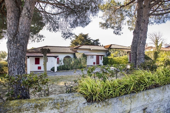 A white building with red shutters and a tiled roof, surrounded by trees and shrubs.