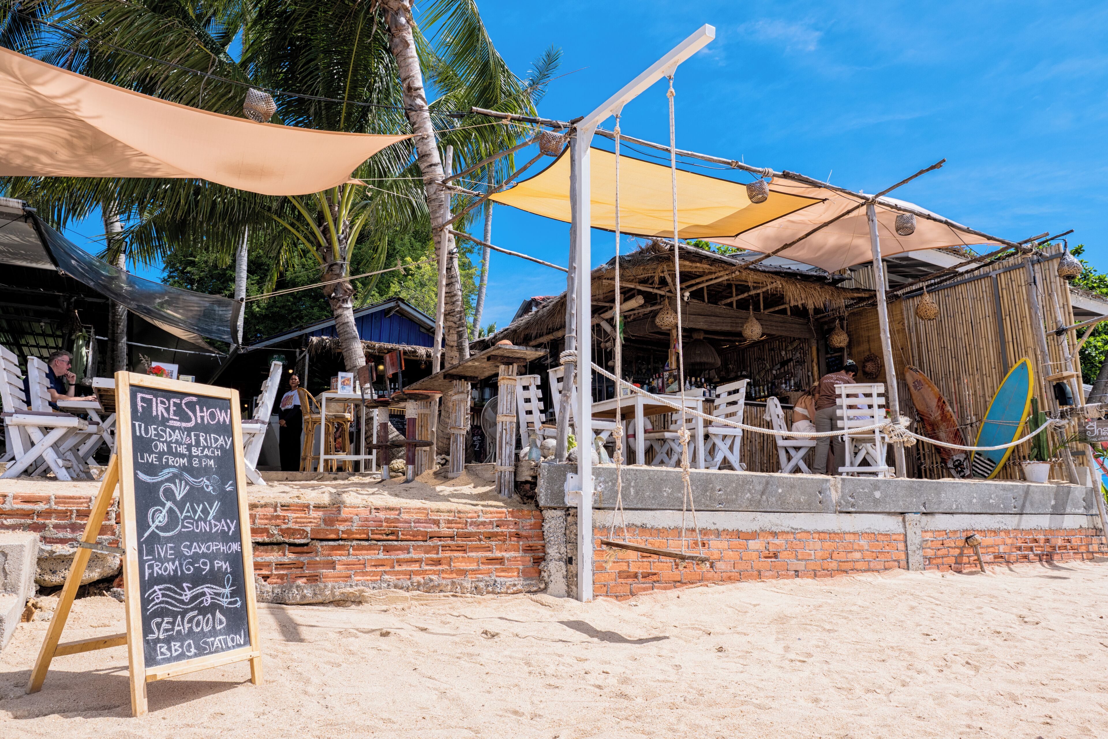 A beachfront restaurant with outdoor seating, a chalkboard sign advertising live saxophone and seafood BBQ, and a surfboard visible in the background.