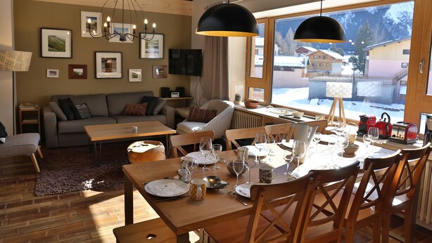 A cozy dining area with wooden furniture, a large window with a view of a snowy landscape, and a chandelier.