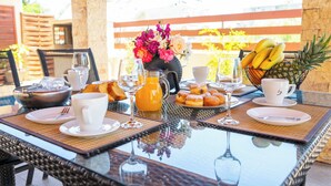 Une table de petit-déjeuner dressée avec des assiettes, des tasses, un pichet de jus d’orange, un saladier de fruits et un panier de bananes et d’ananas.