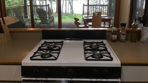 A kitchen with a gas stove, a dining area with a table and chairs, and a view of a forest through large windows. - Seal Point Waterfront Cottage (Lamoine)
