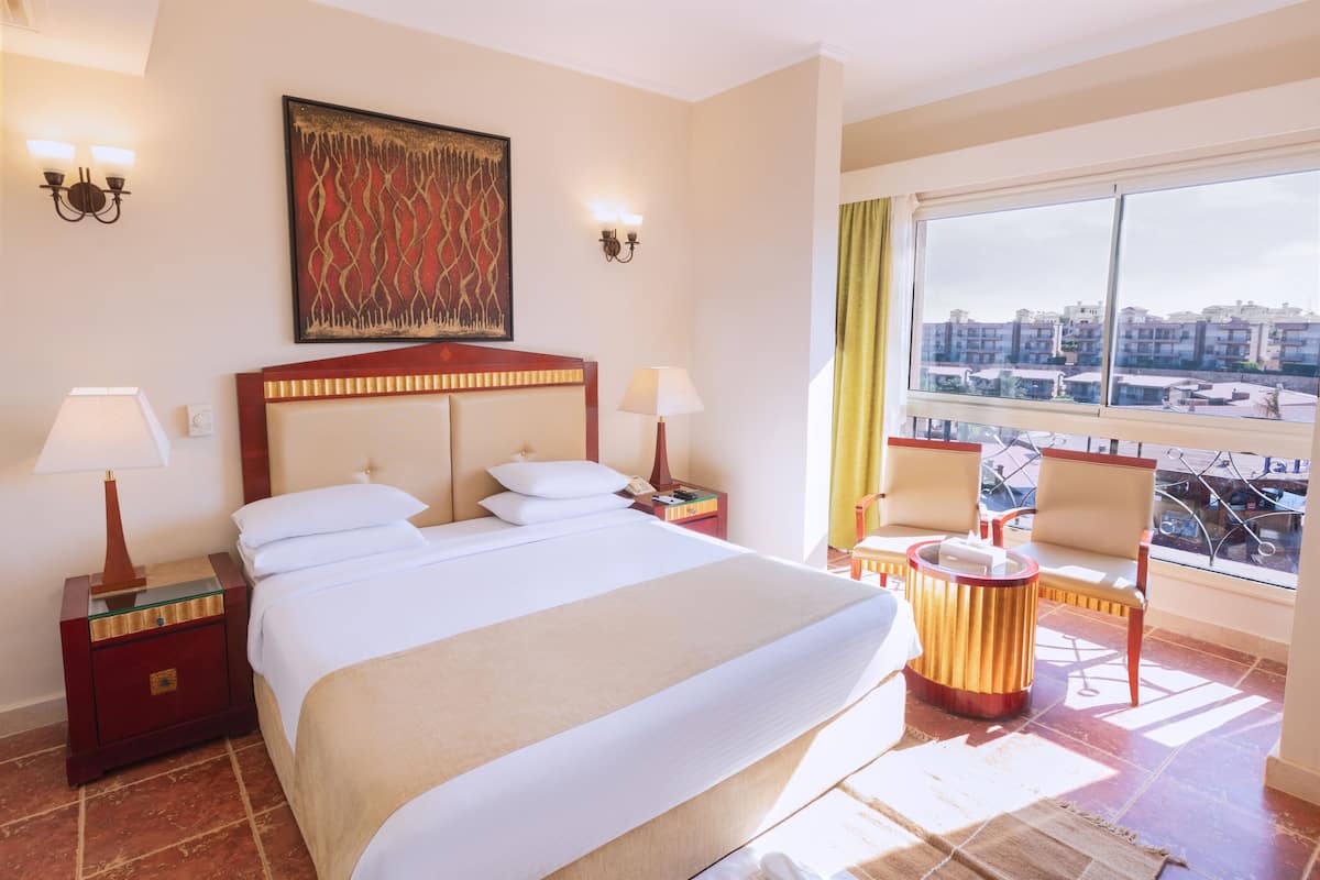 Hotellihuone, jossa on suuri sänky, kaksi yöpöytää valaisimineen, ikkuna, josta avautuu näkymä kaupunkiin, sekä pieni oleskelualue, jossa on tuoli ja pyöreä pöytä.