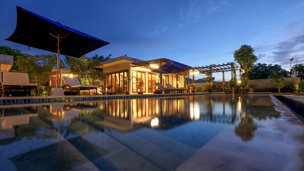 A poolside area with a clear pool, lounge chairs, umbrellas, and a building with large windows and outdoor lighting. - Villa Luna (Kuta)