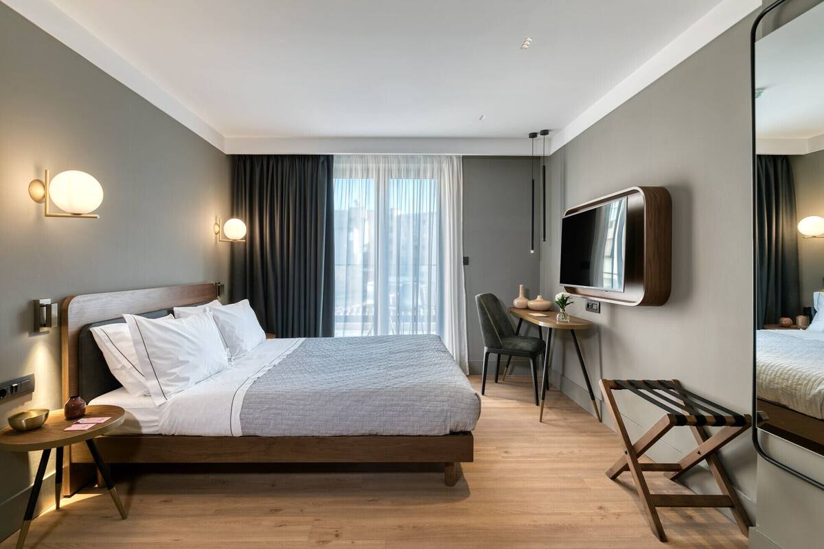 Moderni hotellihuone, jossa on sänky, yöpöytä, tuoli, työpöytä, televisio ja peili.