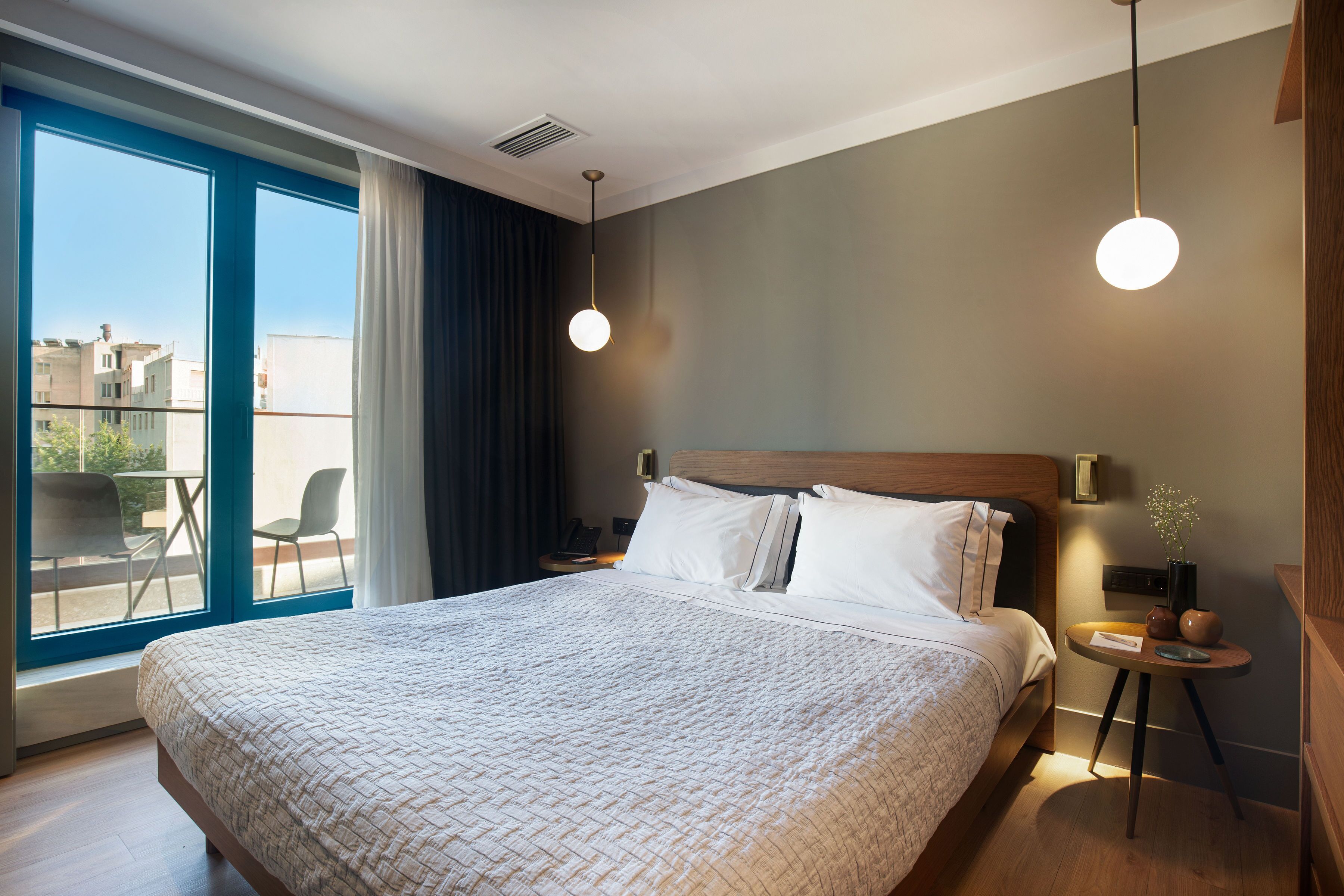 A modern hotel room with a large bed, a bedside table with a lamp, a view of the city through large windows, and a balcony with chairs.
