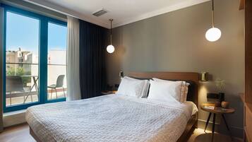 A modern hotel room with a large bed, a bedside table with a lamp, a view of the city through large windows, and a balcony with chairs.