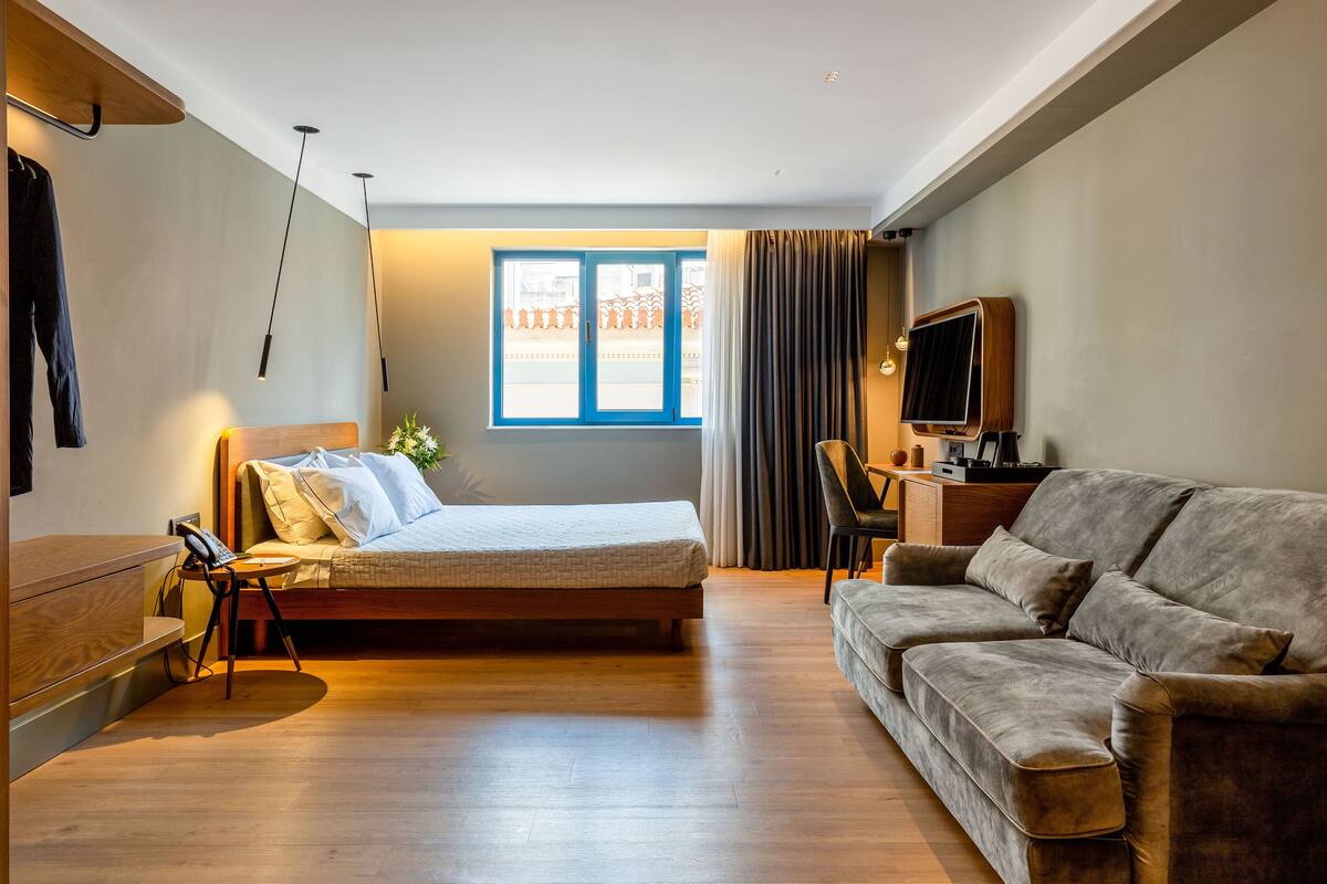 Moderni hotellihuone, jossa on sänky, sohva, työpöytä, jossa on televisio, ja ikkuna, jossa on verhot.