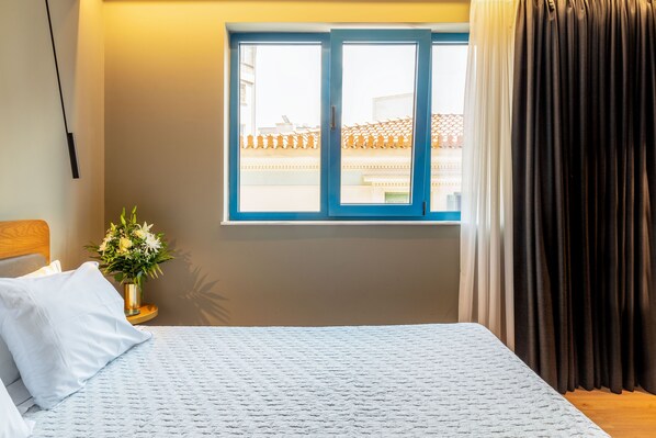 Una habitación de hotel con una cama, una ventana con cortinas, una mesita de noche con un jarrón de flores y vistas a edificios.