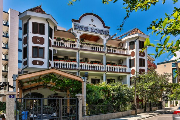 A multi-story building with balconies, a sign reading "MDK Dvor Kornic 1996-1997", and a gated entrance.