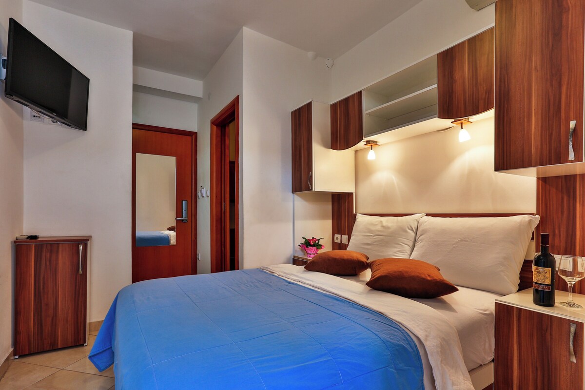 A hotel room with a bed, a TV mounted on the wall, a wooden wardrobe, and a bedside table with a bottle and glass.