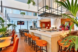 A modern hotel lobby with a high ceiling, multiple seating areas, and a central bar with a large TV screen.