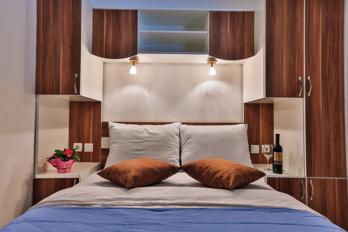 A bedroom with a bed, pillows, a nightstand, a wine bottle, and a vase of flowers.