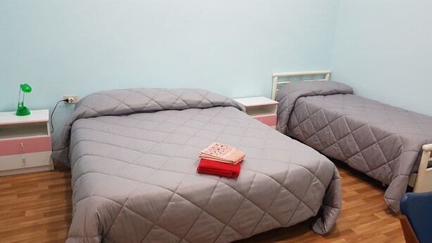 A bedroom with two beds, each with a grey quilted duvet and pillows, a bedside table with a lamp, and a small shelf with a book. - B&B Miraglia (Naples)