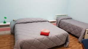 Un dormitorio con dos camas, cada una con cubrecama y almohadas acolchadas de color gris, una mesita de noche con lámpara, y un estante pequeño con un libro.