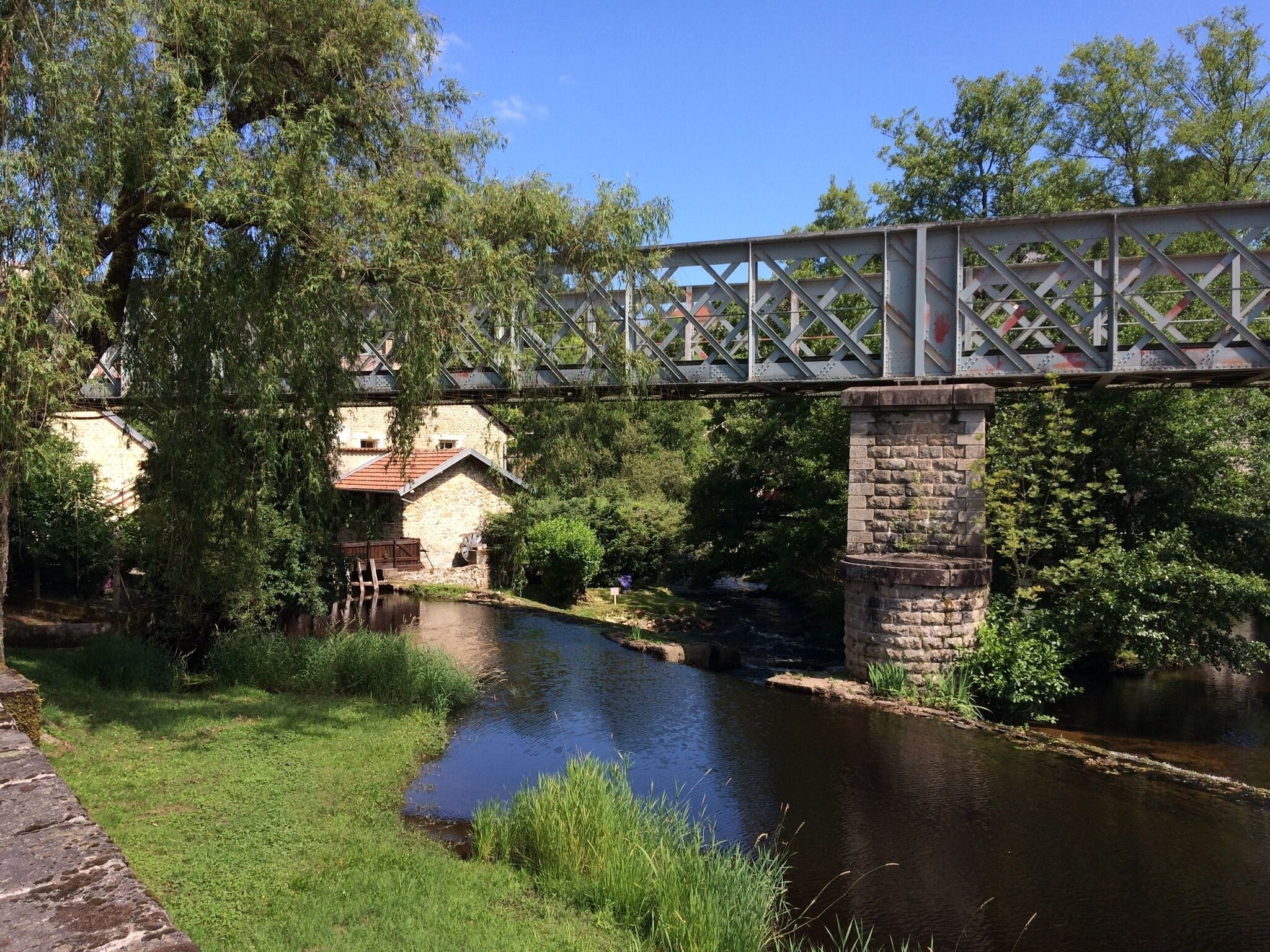 A stone bridge over a calm river, surrounded by lush greenery and a small building with a red roof.