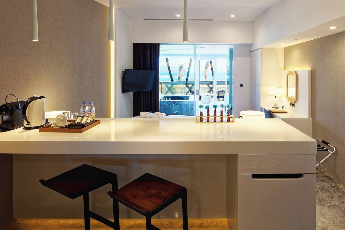 A hotel room with a large central island, a flat-screen TV, a desk with a lamp, and a view of the ocean.